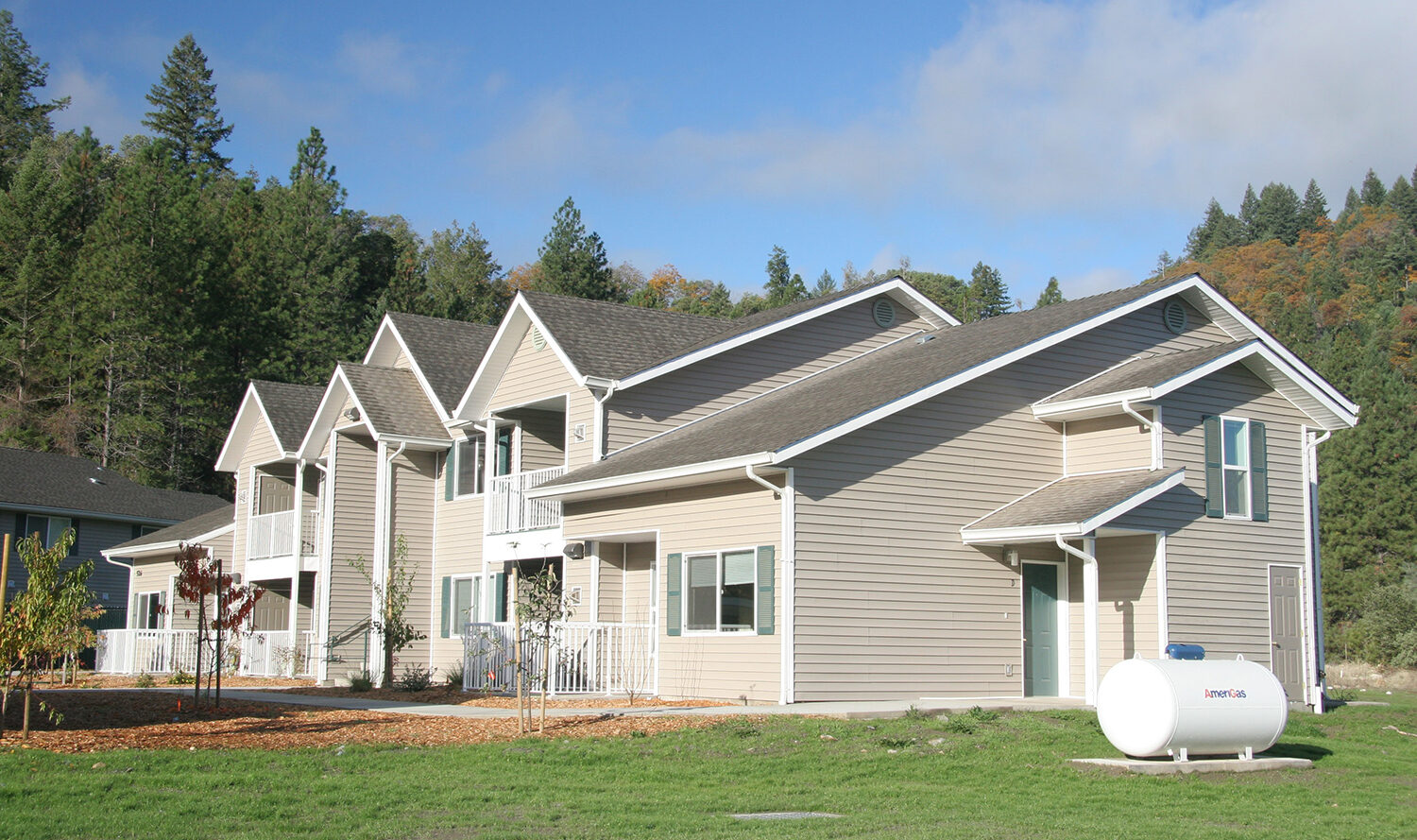 A cluster of two-story beige houses with white trim set against a backdrop of lush green trees and clear blue sky. A propane tank is visible on the grassy lawn in the foreground.