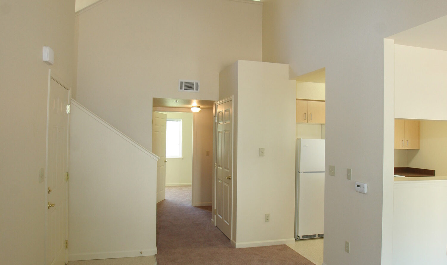 Spacious interior of a vacant, modern apartment with beige walls and carpeting. The room features high ceilings, a staircase, and a glimpse of the kitchen area with white cabinets and refrigerator. Natural light enters through a distant window.