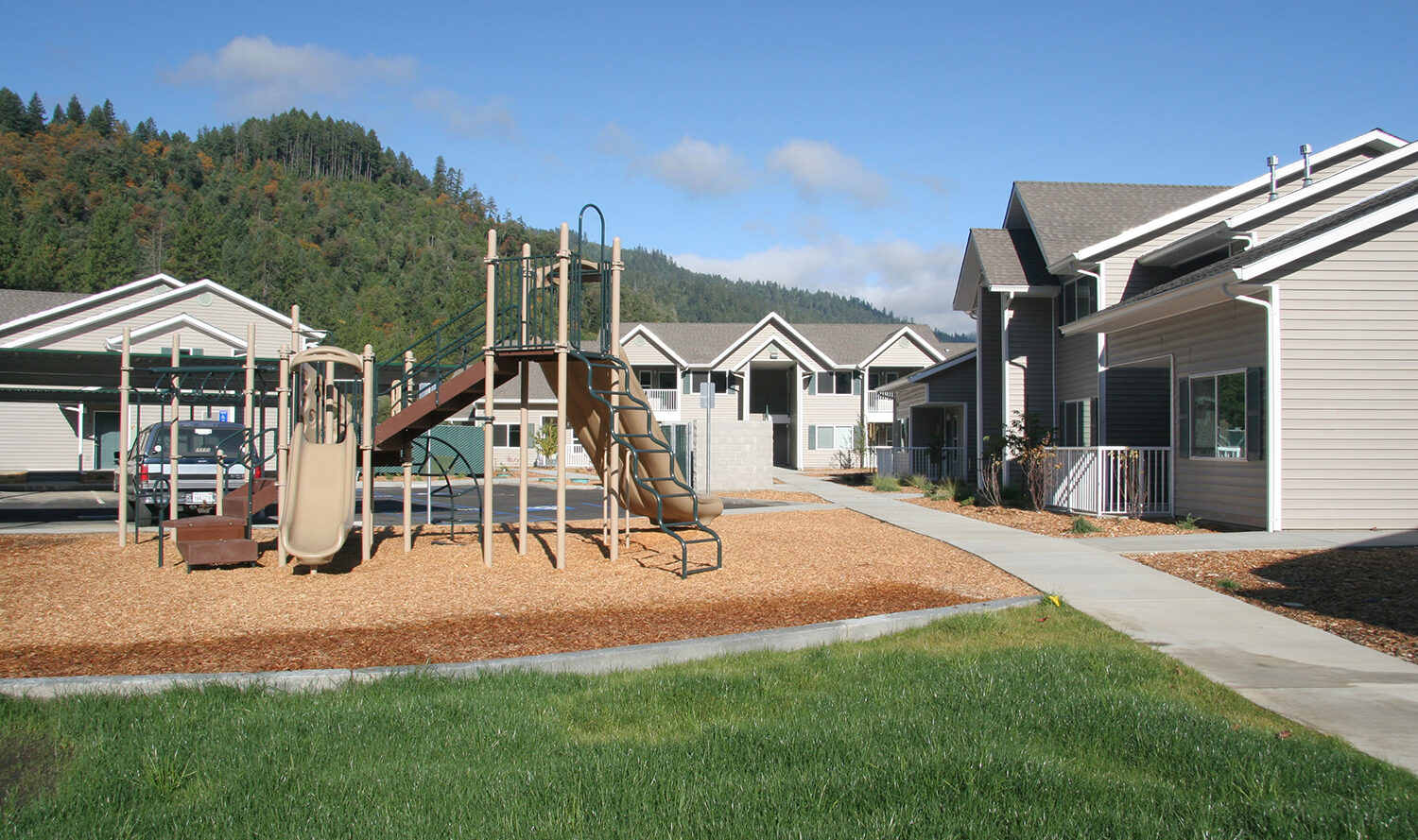 A playground with a slide and climbing structure is surrounded by residential buildings on a sunny day. The area is paved with a grassy lawn in the foreground and wooded hills in the background under a clear blue sky.