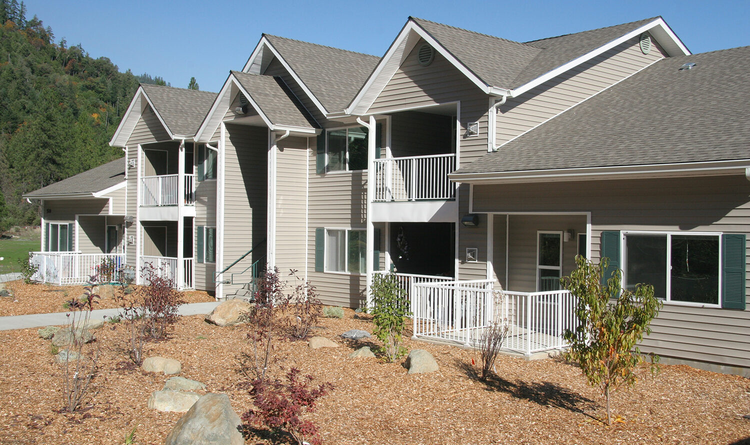 A modern apartment building with beige siding and white trim, featuring multiple peaked roofs and small balconies. The landscaping includes neatly arranged rocks and young shrubs, set against a backdrop of green, forested hills under a clear blue sky.