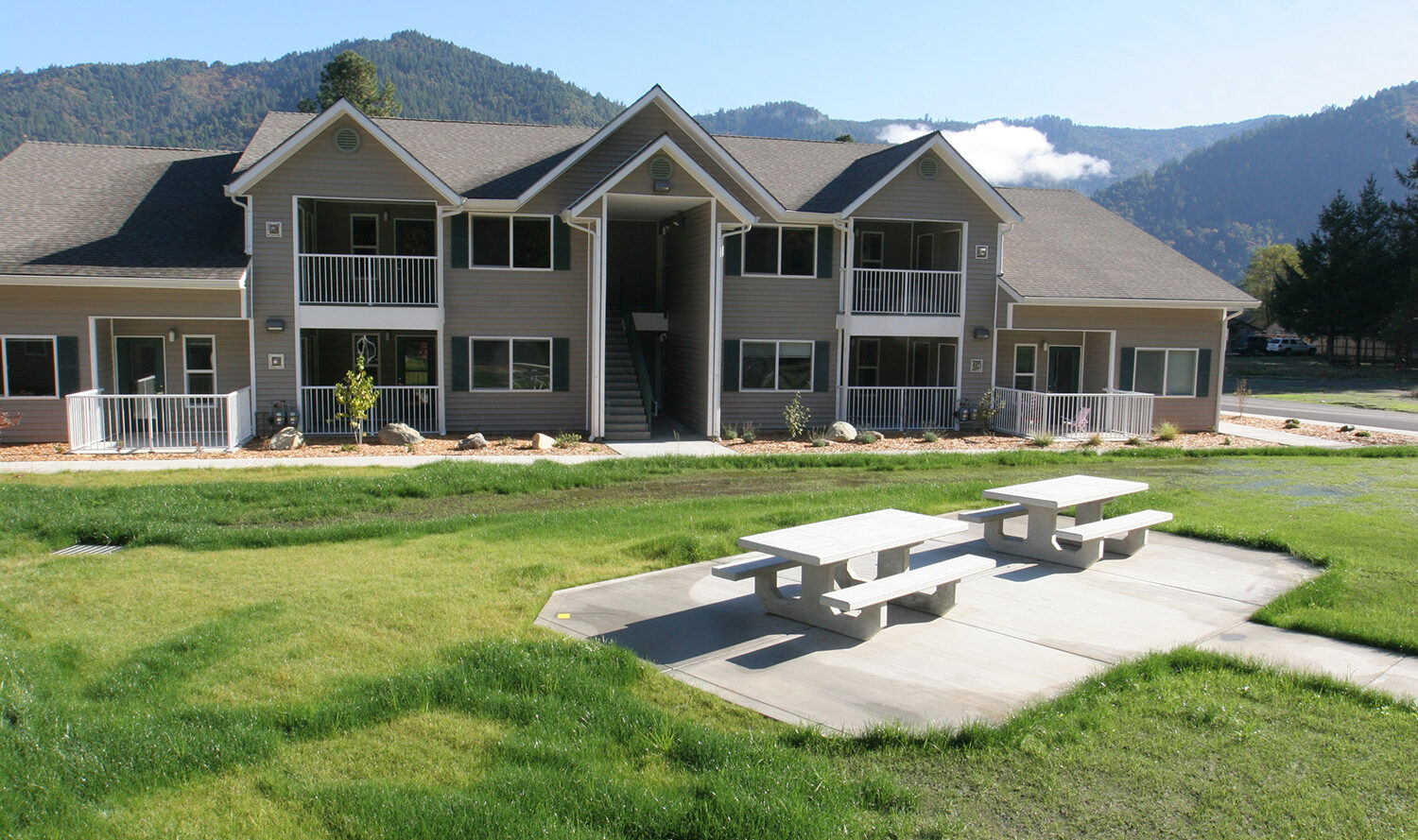 A two-story gray apartment building with balconies, nestled among green grass and trees. Two white picnic tables are on the concrete patio in front. Hills and a clear sky serve as the backdrop.