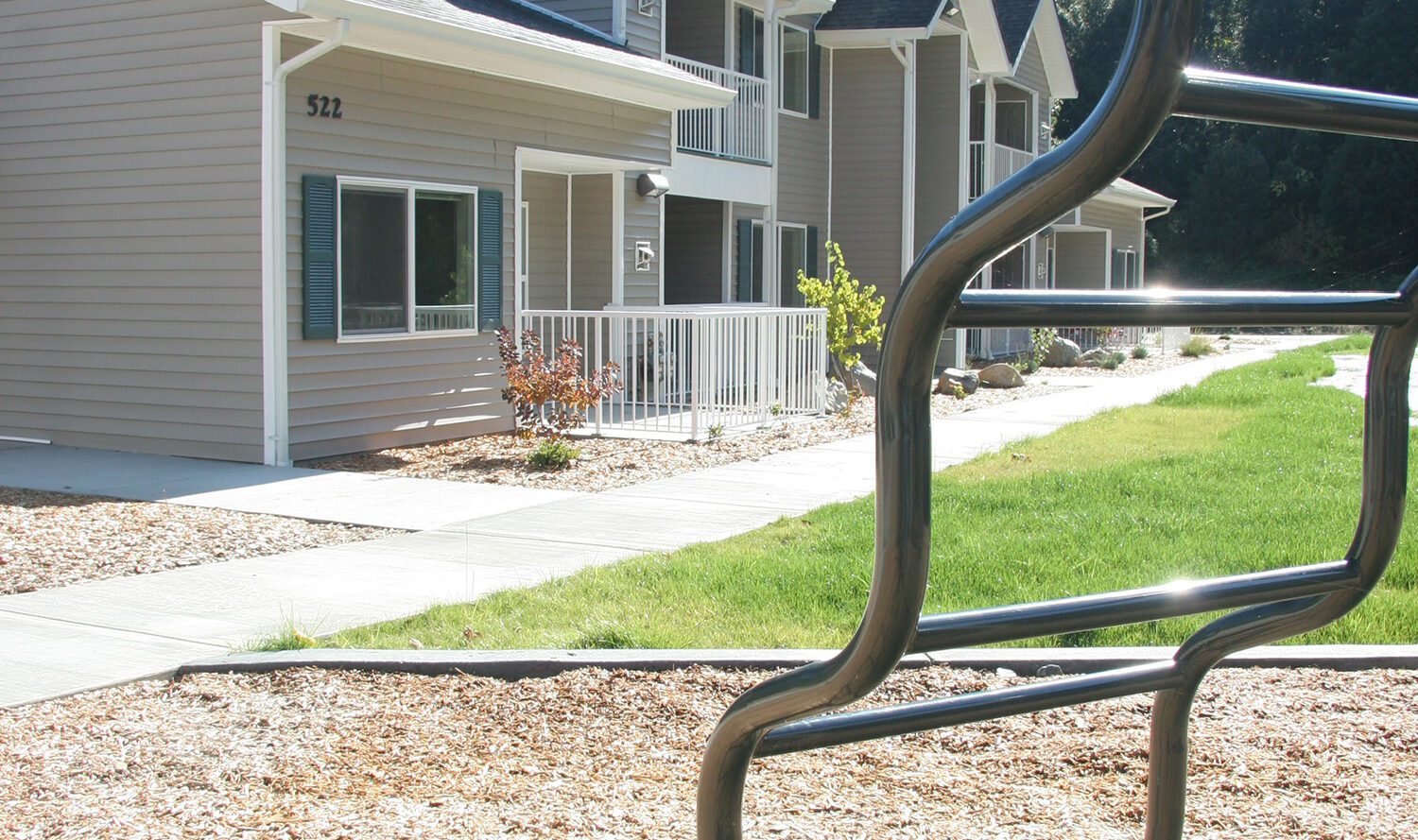 A playground climbing structure stands in front of a two-story residential building with balconies. The area is surrounded by greenery and trees under a clear blue sky.