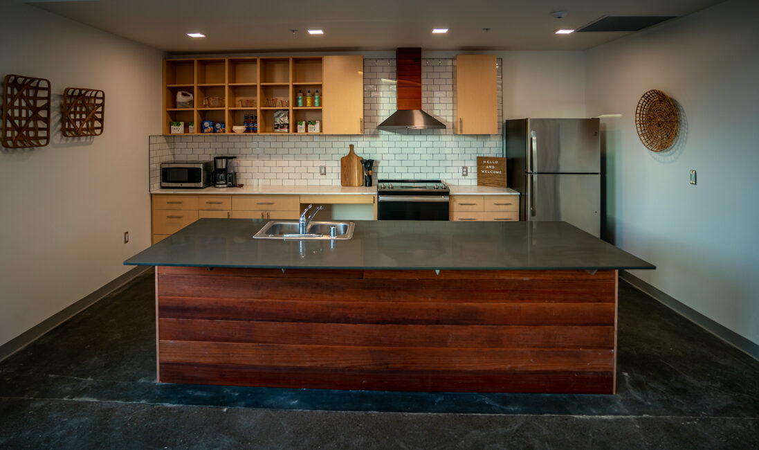 A modern kitchen with a central island featuring a dark countertop. Behind it are light wooden cabinets, white subway tile backsplash, stainless steel appliances, and decorative wall hangings. The space is well-lit and minimalistic.