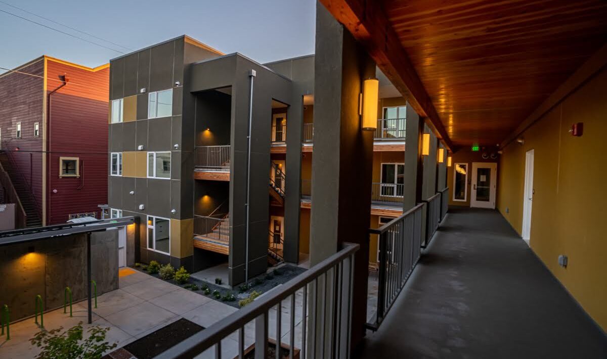 View of a modern apartment complex from an upper-level walkway, showing multiple units with large windows. The setting is during twilight, with warm outdoor lighting and a landscaped courtyard below.