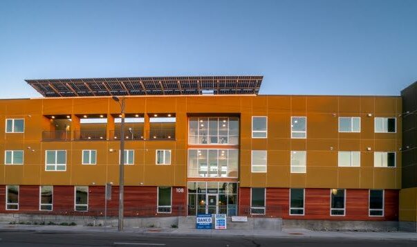 A modern, three-story building with a geometric facade features large windows and a mix of yellow, orange, and brown panels. Solar panels are visible on the roof. The building is situated along a street under a clear blue sky.