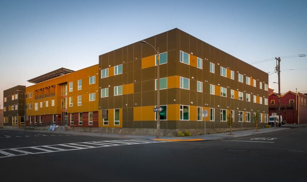 A modern, angular building with a mix of yellow and brown panels against a clear sky. The structure has large windows and occupies a street corner, with a crosswalk and street signs visible in the foreground.