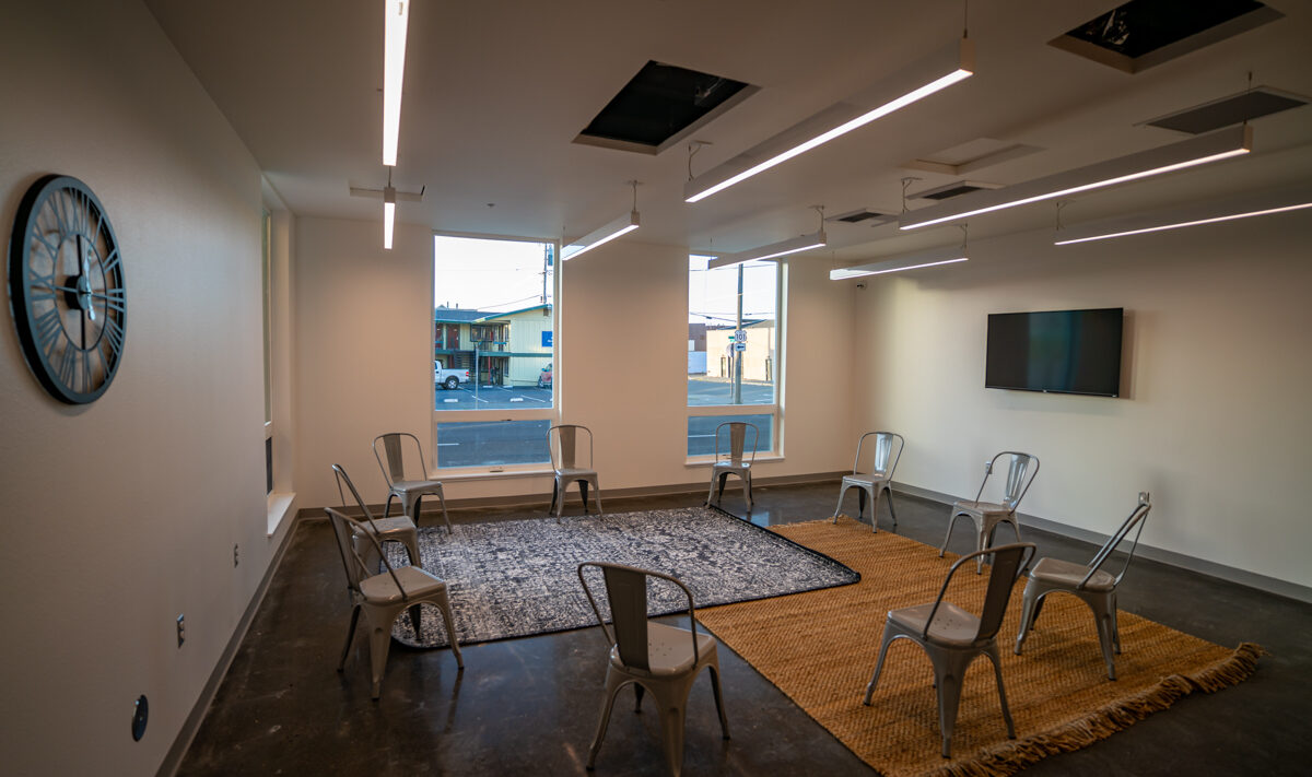 A meeting room with metal chairs arranged in a circle on two rugs, one black and one tan. A clock is on the left wall, and a flat-screen TV is mounted on the right wall. Large windows provide natural light. Ceiling lights are turned on.