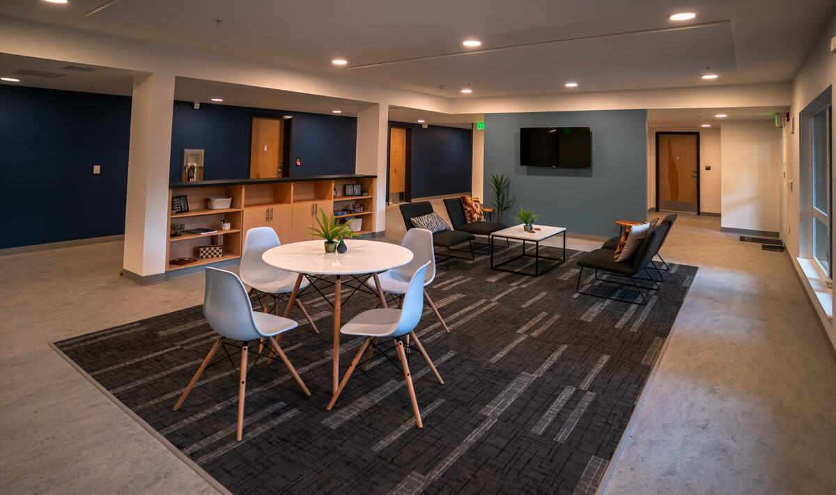 A modern lobby with a seating area featuring black couches, a round white table with chairs, and ornamental plants. The room has blue accent walls, a mounted TV, and open shelving with decorative items.