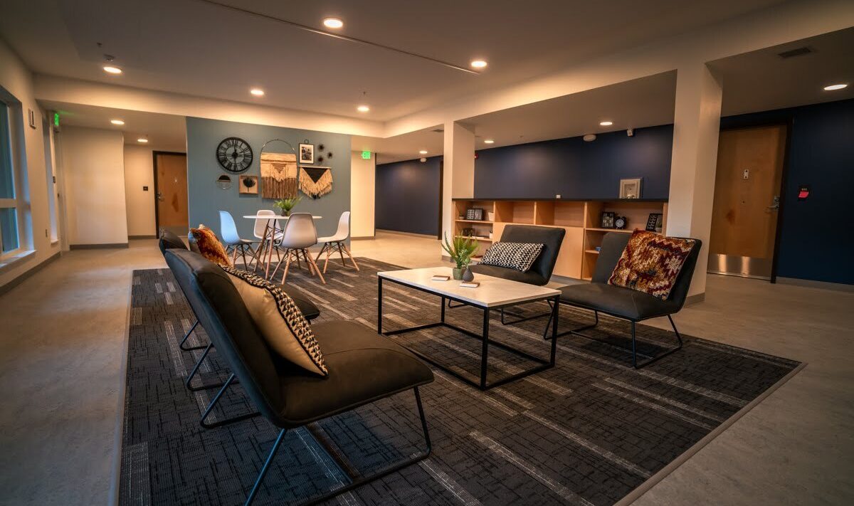 A modern lounge area with black couches and a rectangular coffee table on a dark patterned rug. In the background, theres a dining table with white chairs, decorative wall items, and shelves against a blue accent wall.