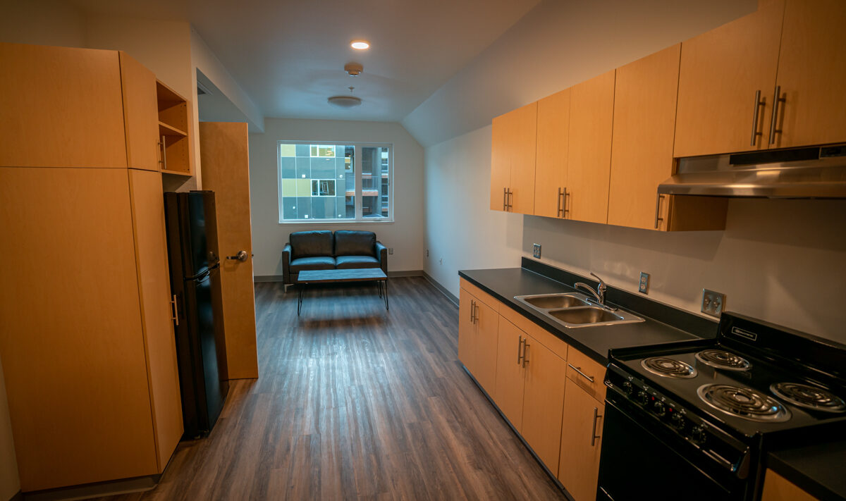 A modern apartment interior with wooden floors features a compact kitchen on the right with light wood cabinets, a black countertop, sink, stove, and fridge. A small living area with a gray couch is in the background near a window.