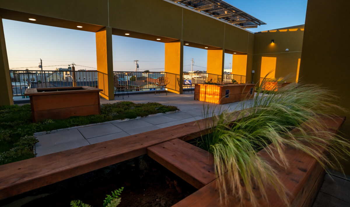 A rooftop garden with wooden planters filled with greenery, surrounded by yellow walls and open archways. Solar panels are visible above, and city buildings are seen in the background against a clear blue sky.