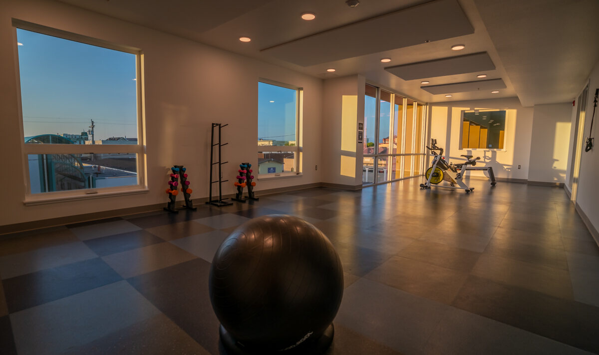 A gym room with large windows showcasing a scenic view. The room features an exercise ball, a small rack of colorful dumbbells, a stationary bike, and a set of weights. Warm lighting fills the space, creating a serene exercise environment.