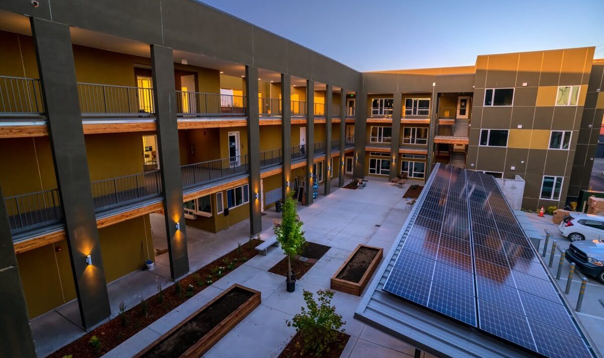 A modern apartment complex with multiple floors and balconies arranged around a central courtyard. Solar panels are installed on a carport, and there are planter boxes with small trees in the courtyard. The sky is clear, suggesting it is evening or early morning.