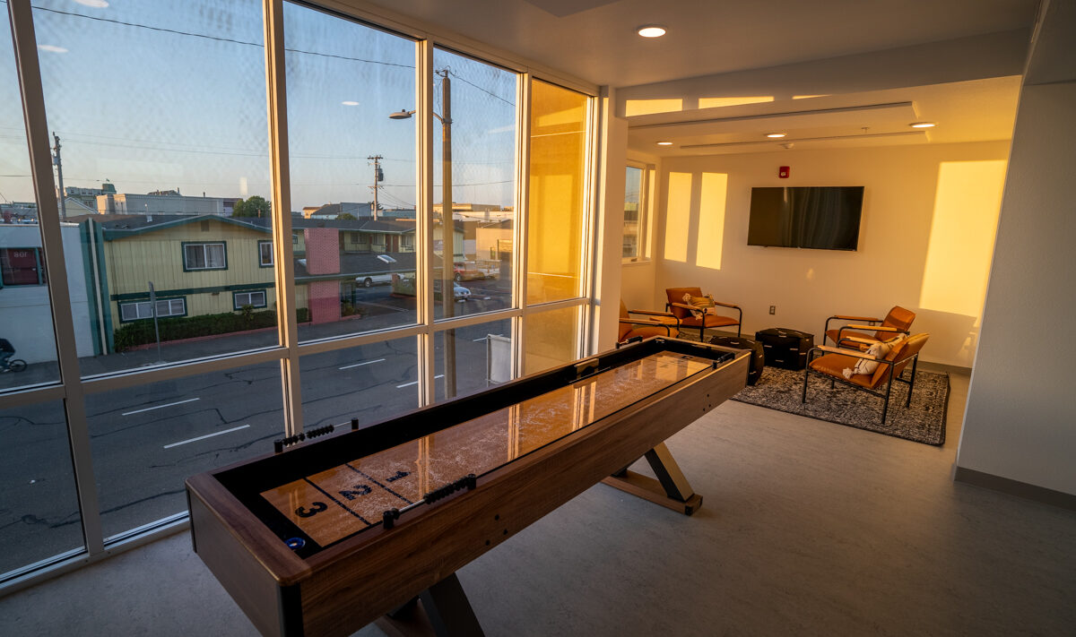 A brightly lit room with large windows showcasing a shuffleboard table. Two modern chairs and a small table sit in front of a wall-mounted TV. Outside, a street and colorful houses are visible against a clear sky.