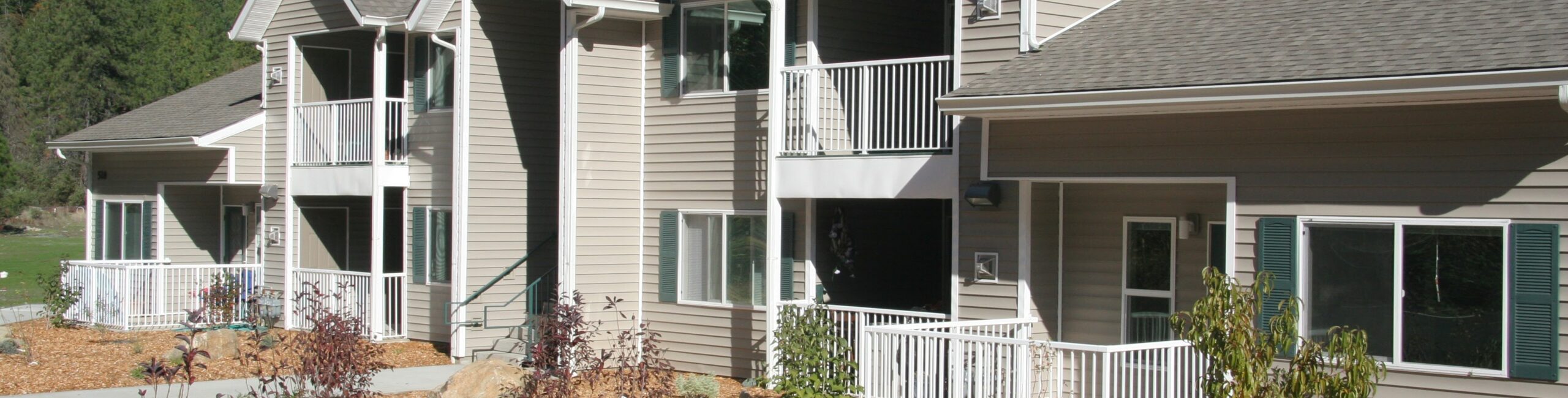 A row of contemporary two-story apartment buildings with beige siding and white trim. Each unit has a small balcony or porch. The landscaped area features mulch, small bushes, and rocks. Tall trees and a clear blue sky are in the background.