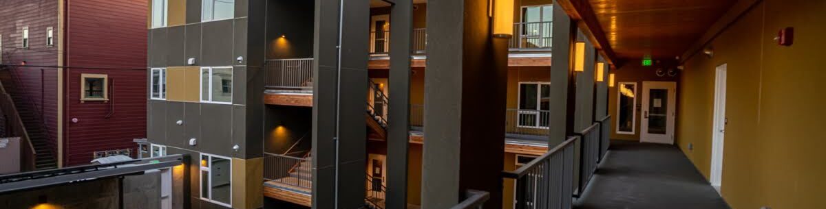 View of a modern apartment complex from an upper floor walkway. The building features dark green and maroon exteriors with large windows. The corridor has a wooden ceiling and is lit by warm wall lights.