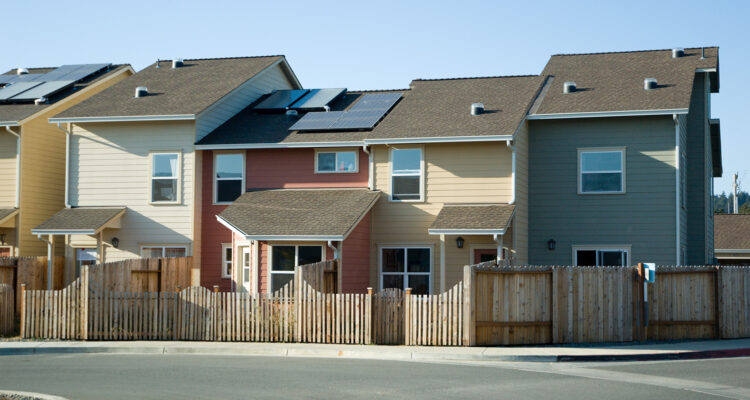 A row of modern townhouses in pastel colors, with a wooden fence in front. The houses feature solar panels on their roofs under a clear blue sky. The view is from a street with a curb in the foreground.