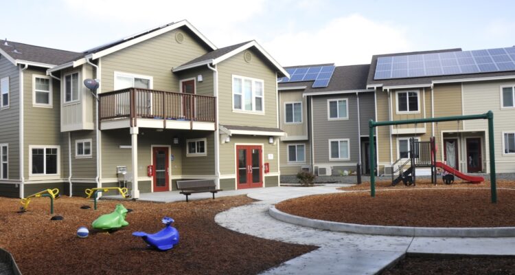 A residential complex with solar panels on the roofs, featuring a playground with colorful equipment, including a slide and rocking toys, set on a mulch surface. A curved concrete path leads around the play area.