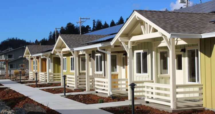 A row of small, colorful houses with front porches line a sidewalk under a clear blue sky. The houses have white trim and solar panels on the roofs. Mulched garden beds border the walkway.