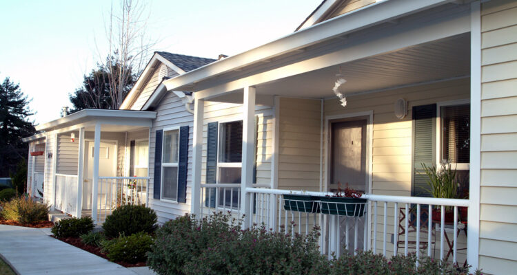 A row of single-story houses with front porches and beige siding. The houses have white trim and are bordered by small shrubs. A sidewalk runs along the front of the homes, with trees visible in the background.