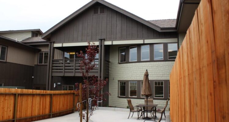 A two-story house with a wooden exterior and a sloped roof. The patio features a table, chairs, and an umbrella. A wooden fence surrounds the area, and theres a small tree in the concrete yard.